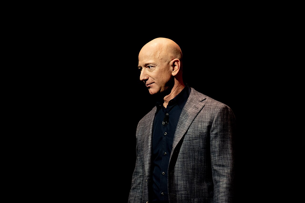 Jeff Bezos de paletó cinza e camisa escura, visto de perfil, iluminado em um palco escuro durante um evento da Blue Origin.