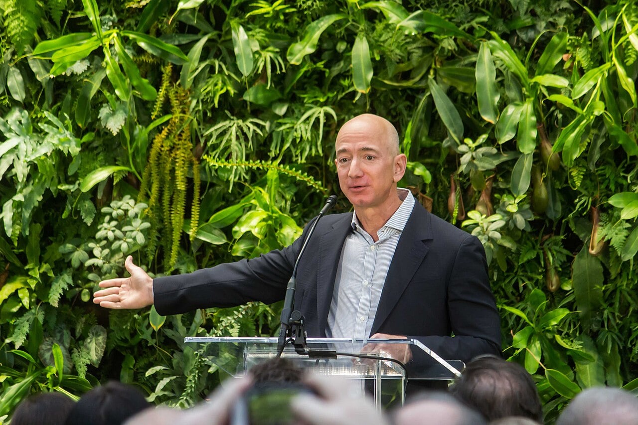 Jeff Bezos, de paletó escuro, discursa ao microfone em um púlpito de acrílico, diante de uma parede de plantas, com a plateia em primeiro plano.