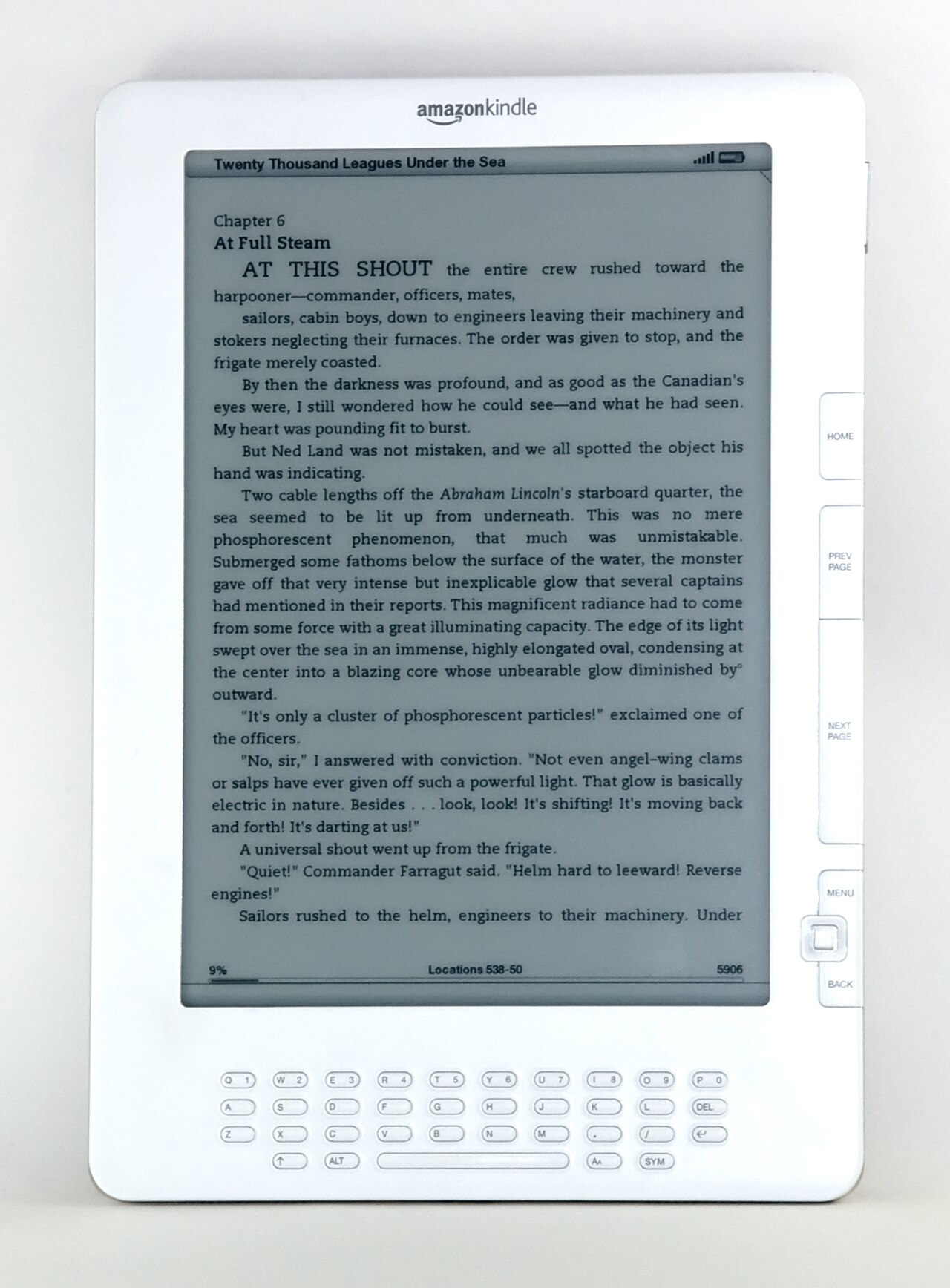 Leitor Kindle DX branco, visto de frente, com tela de tinta eletrônica exibindo um trecho de livro em inglês e teclado físico abaixo.
