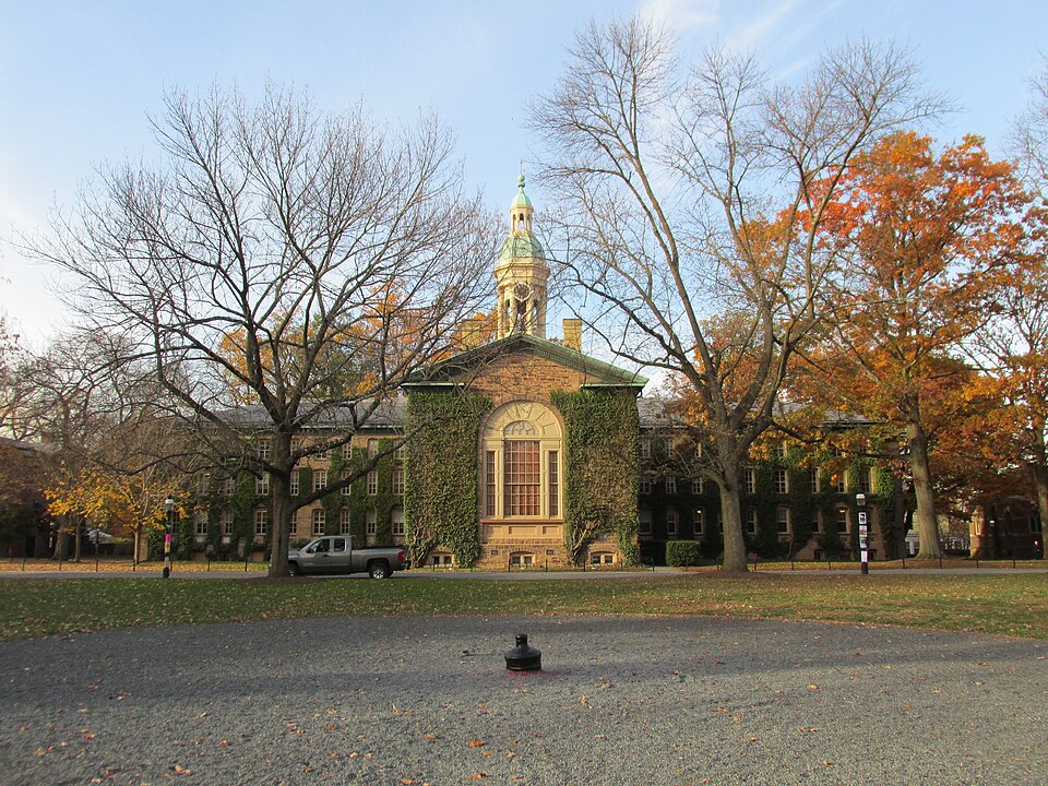 Fachada de tijolos e pedra de Nassau Hall, coberta de hera, com uma torre de relógio no centro, árvores de outono e um gramado à frente.