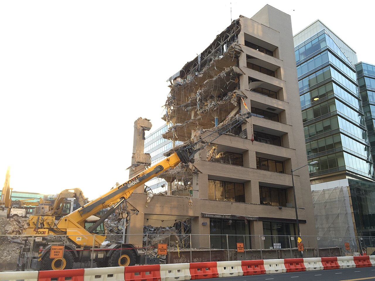 Prédio de escritórios de concreto parcialmente demolido, com andares abertos e uma grua amarela em primeiro plano, ao lado de um edifício de vidro.