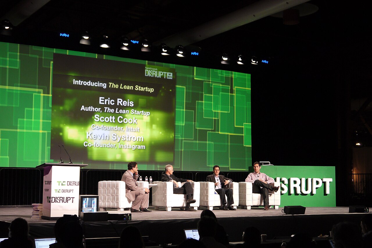Quatro pessoas sentadas em poltronas brancas num palco do TechCrunch Disrupt, diante de um telão que lista Eric Ries, Scott Cook e Kevin Systrom.