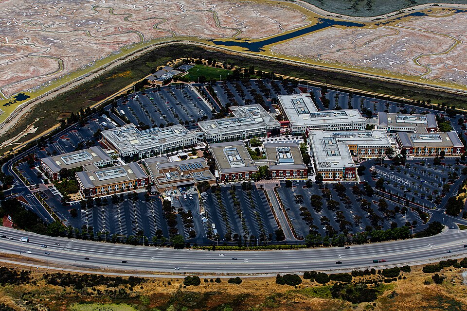 Vista aérea de prédios de escritórios do Facebook em Menlo Park, cercados por estacionamentos, uma rodovia e áreas alagadas da baía.
