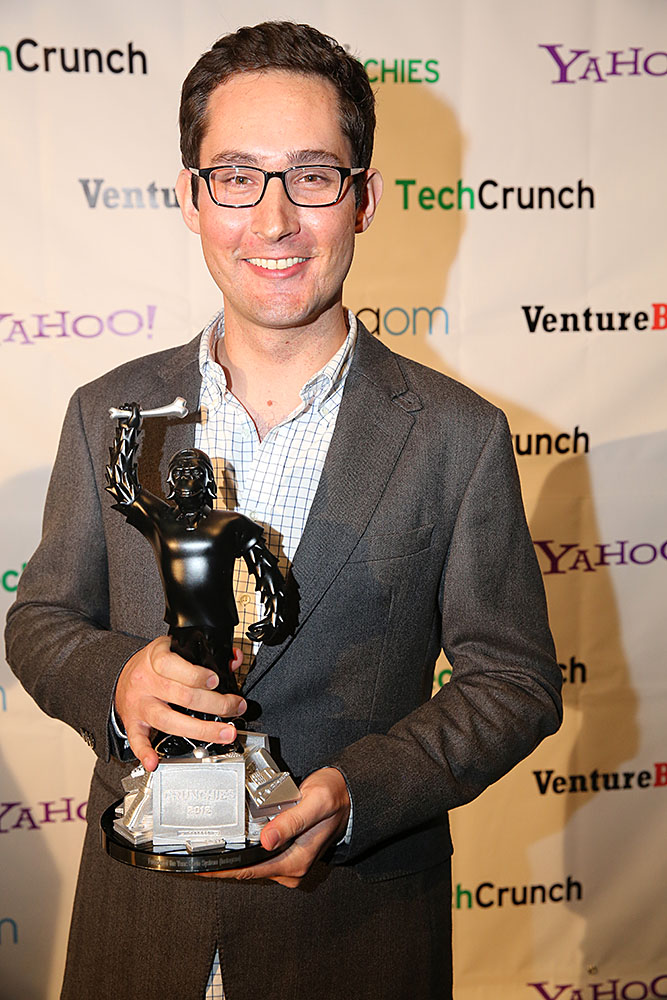 Kevin Systrom de óculos e paletó cinza, sorrindo e segurando um troféu preto, diante de um painel com logotipos de patrocinadores.