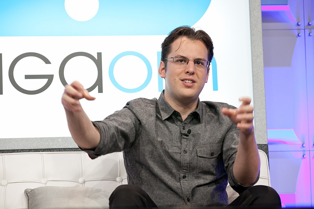 Mike Krieger gesticulando com as duas mãos durante uma conversa, sentado num sofá branco, com um painel claro ao fundo.