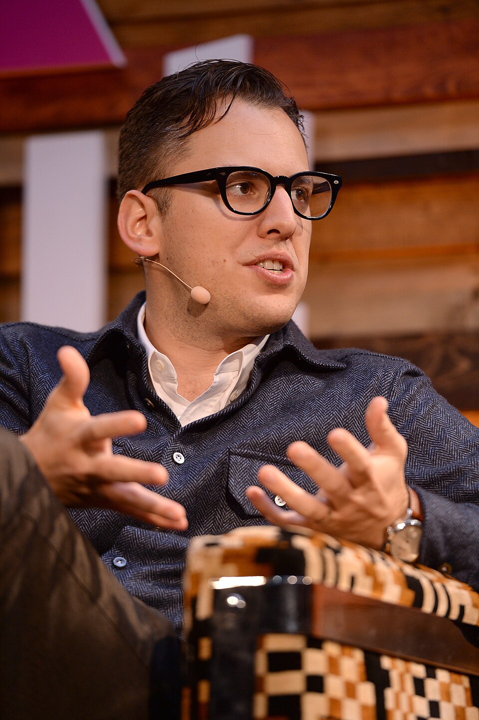 Mike Krieger de óculos de armação preta e camisa azul, gesticulando durante uma conversa, com microfone de cabeça e fundo de madeira.
