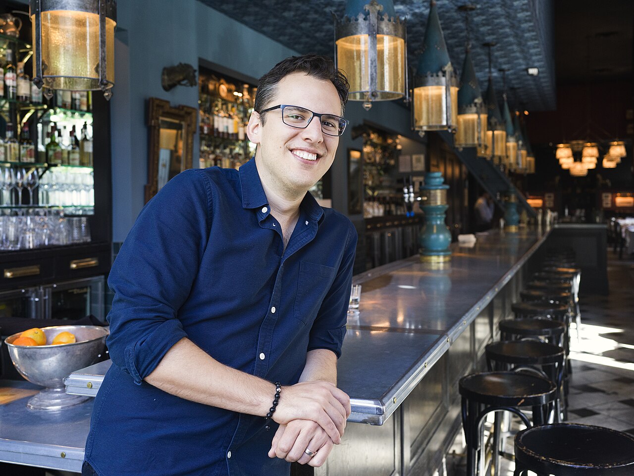 Mike Krieger de camisa azul e óculos, sorrindo, apoiado numa bancada de bar com banquetas e luminárias ao fundo.