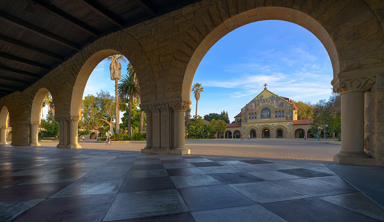 Arcadas de arenito do campus da Universidade Stanford, com a fachada da Igreja Memorial ao fundo, num dia de céu claro.