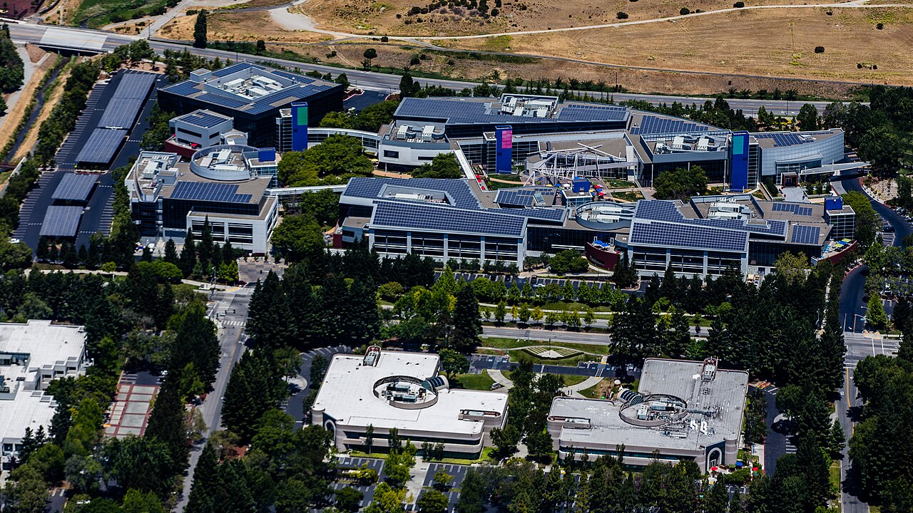 Vista aérea do campus do Google em Mountain View, com prédios de vidro e painéis solares nos telhados, cercados de árvores e estacionamentos.