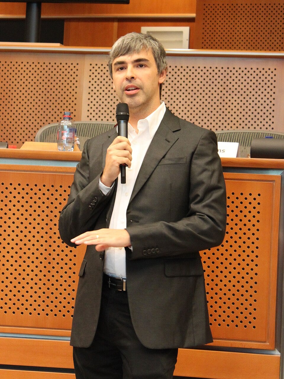 Larry Page em pé, de paletó escuro e camisa branca, falando ao microfone num plenário com painéis de madeira ao fundo.