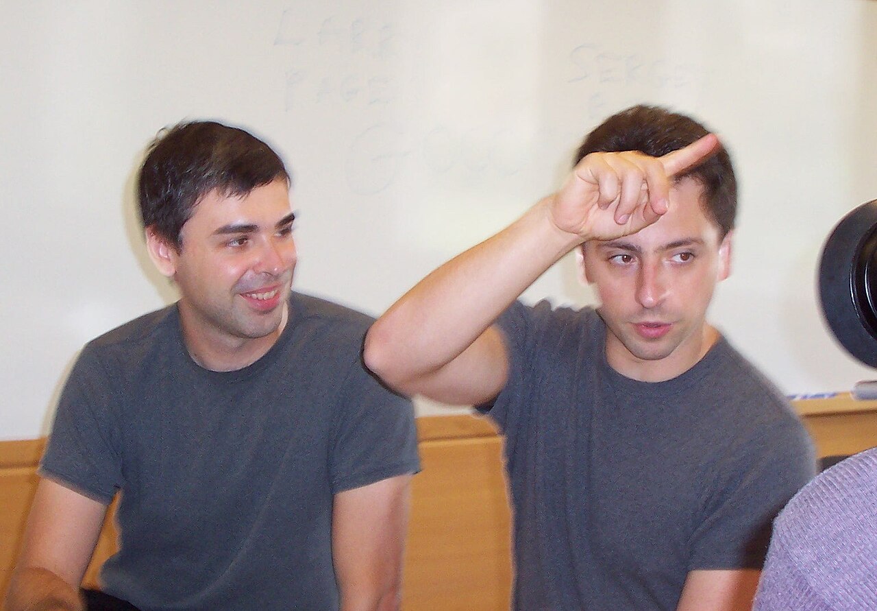 Dois homens de camiseta cinza sentados diante de um quadro branco: um sorri e o outro levanta a mão perto da testa enquanto fala.