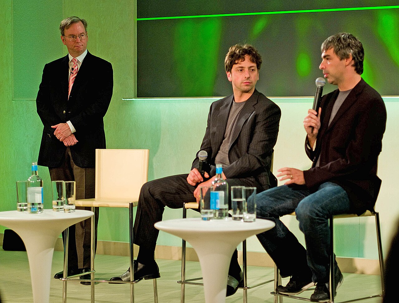 Eric Schmidt em pé à esquerda; Sergey Brin e Larry Page sentados em banquetas, com microfones, num palco com iluminação verde.