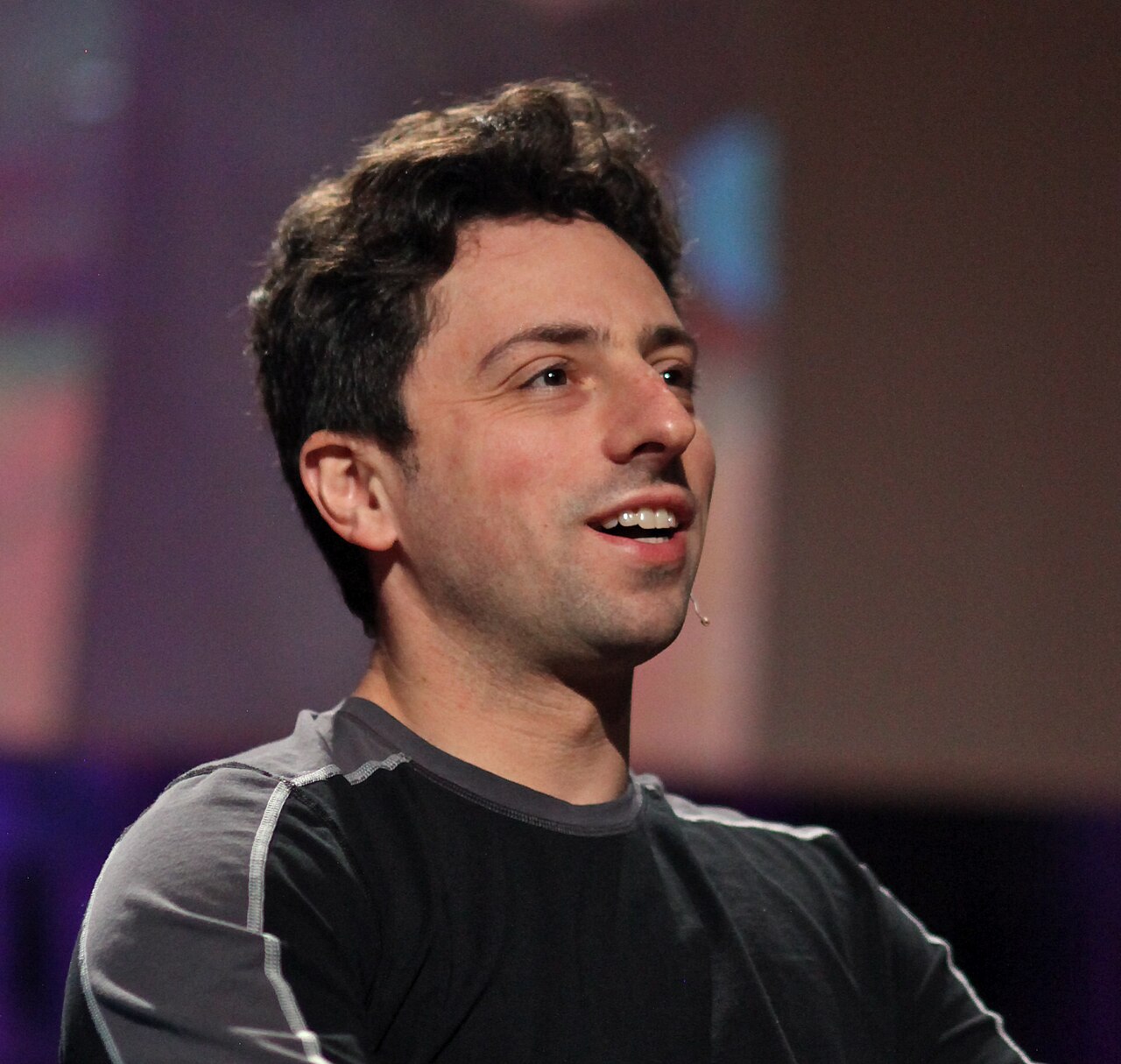 Sergey Brin de perfil, sorrindo, com microfone de cabeça preso à bochecha, num palco com fundo desfocado em tons de roxo e rosa.