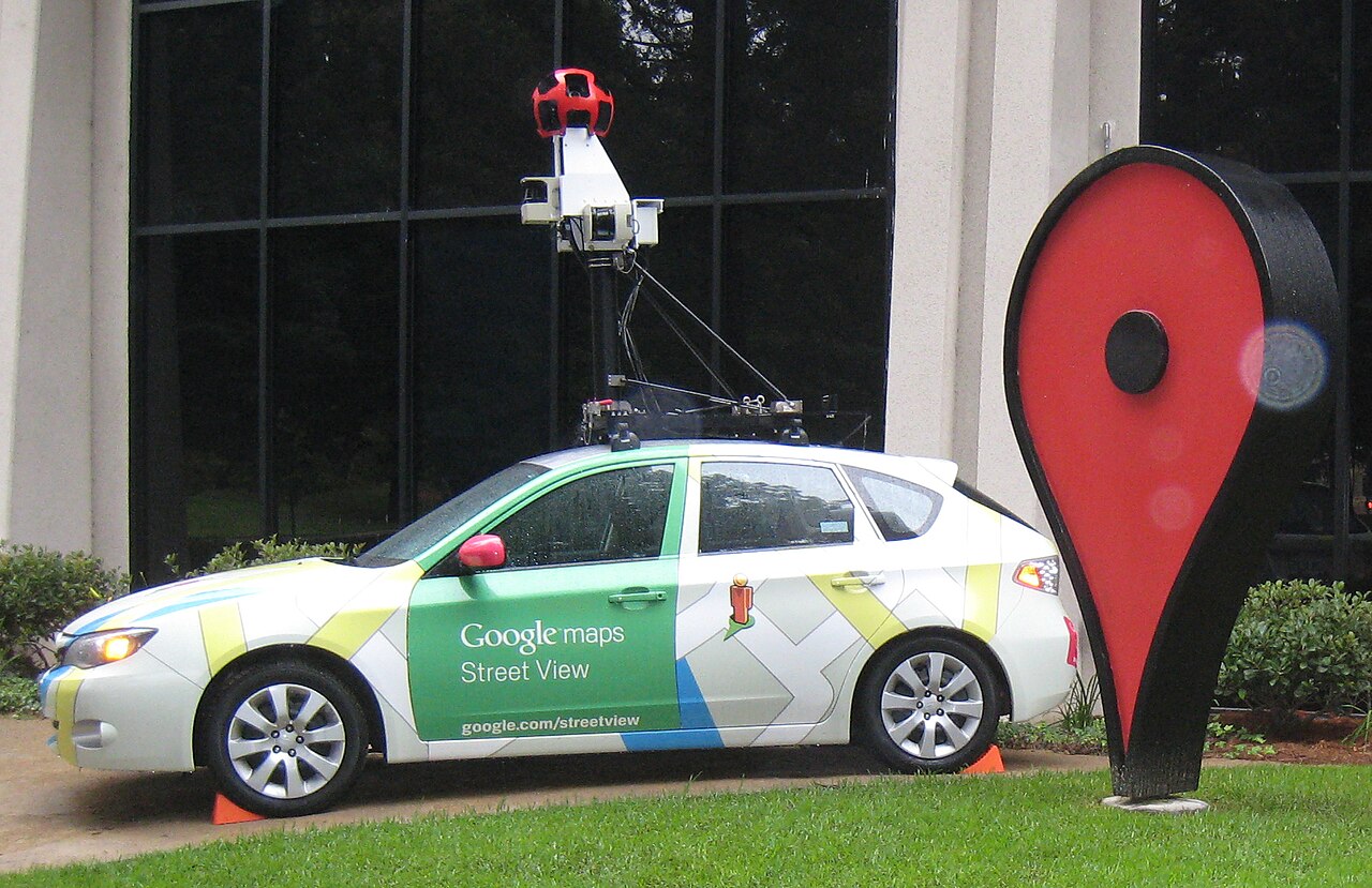 Carro hatch branco e verde, com a inscrição Google Maps Street View, câmera sobre um mastro no teto e um grande marcador de mapa vermelho ao lado.