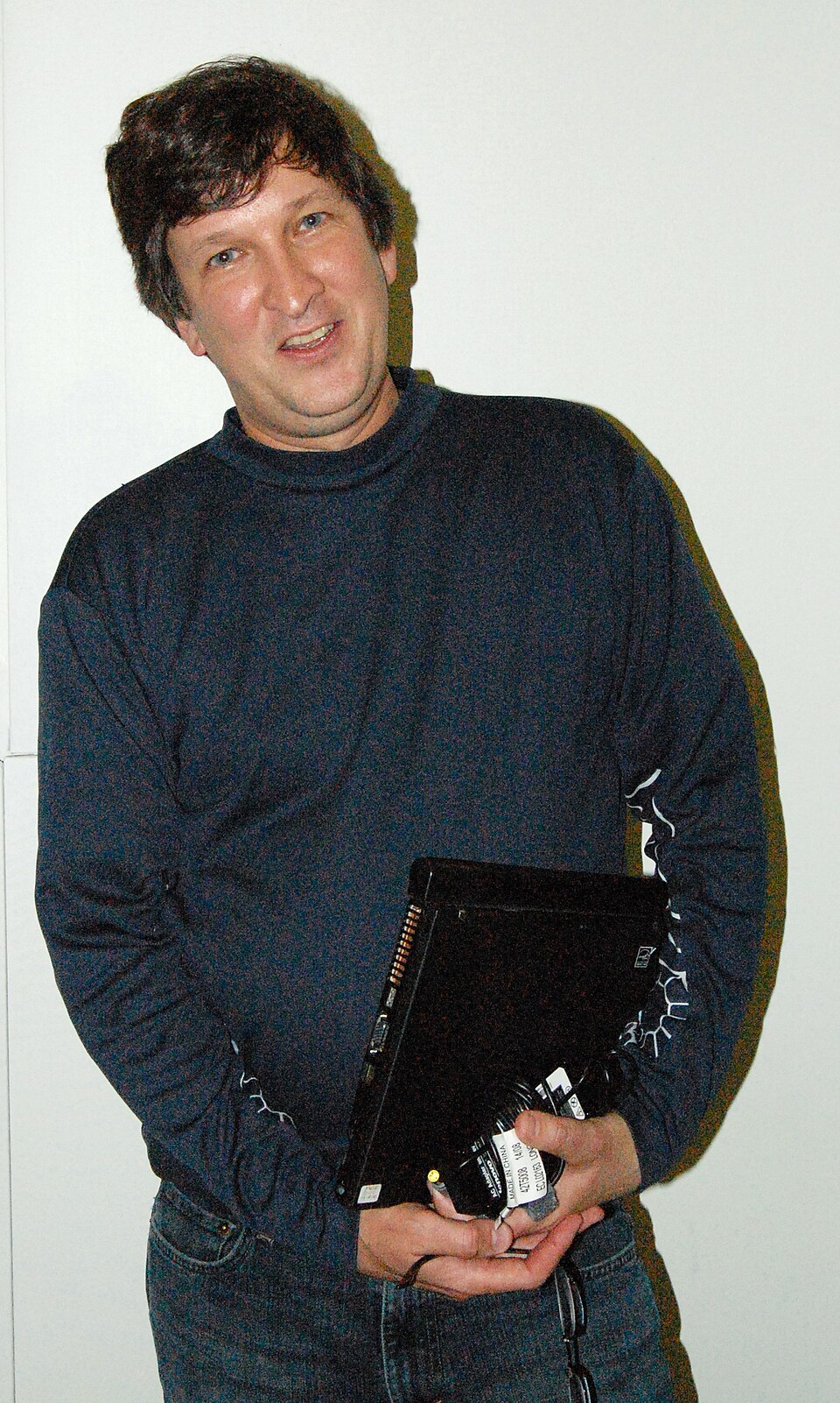 Larry McVoy, de cabelo escuro e blusa de gola alta azul-marinho, segurando um notebook fechado sob o braço, sorrindo diante de uma parede clara.