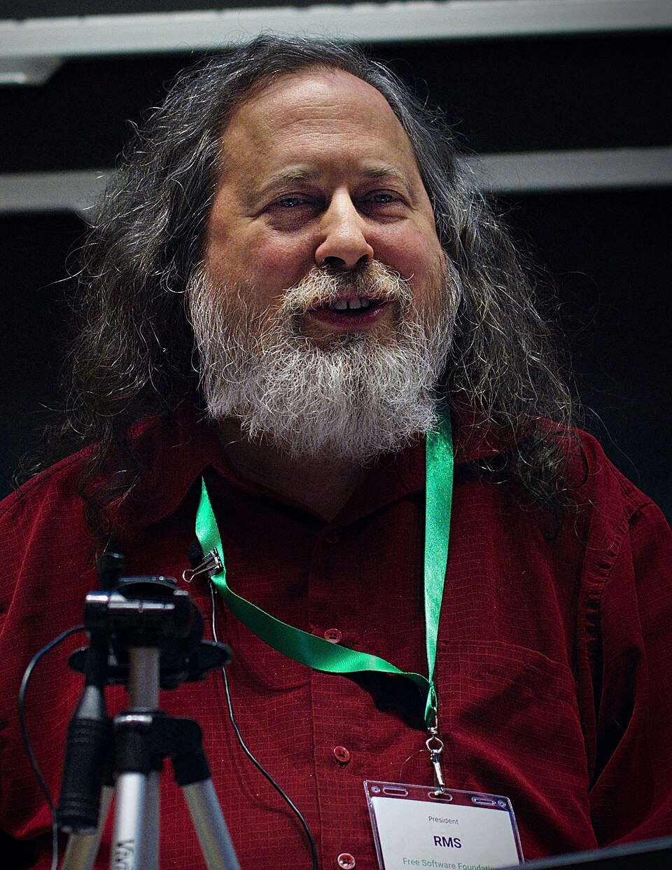 Retrato de Richard Stallman, de cabelos longos grisalhos e barba, camisa vermelha e crachá com a sigla RMS, diante de um tripé.