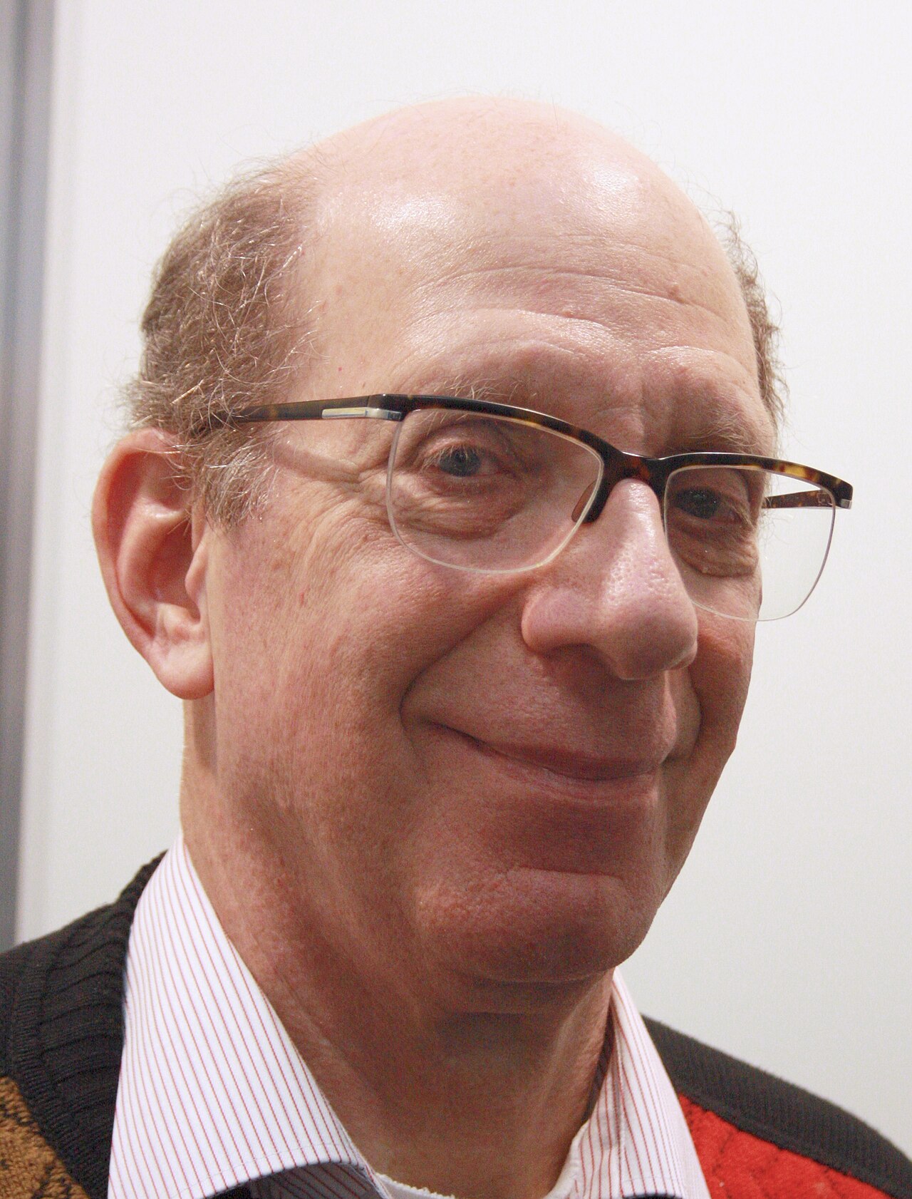 Retrato de Andrew Tanenbaum, homem calvo de óculos, camisa listrada e suéter escuro, sorrindo de lado.