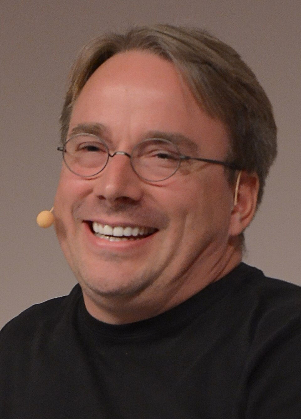 Rosto de Linus Torvalds sorrindo de olhos semicerrados, de óculos redondos e fone de microfone, em blusa preta.