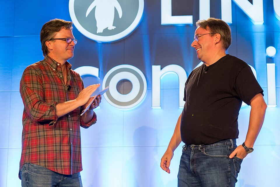 Dois homens de óculos num palco azul do LinuxCon: um de camisa xadrez, aplaudindo; outro, de camiseta preta e mão na cintura, sorrindo.
