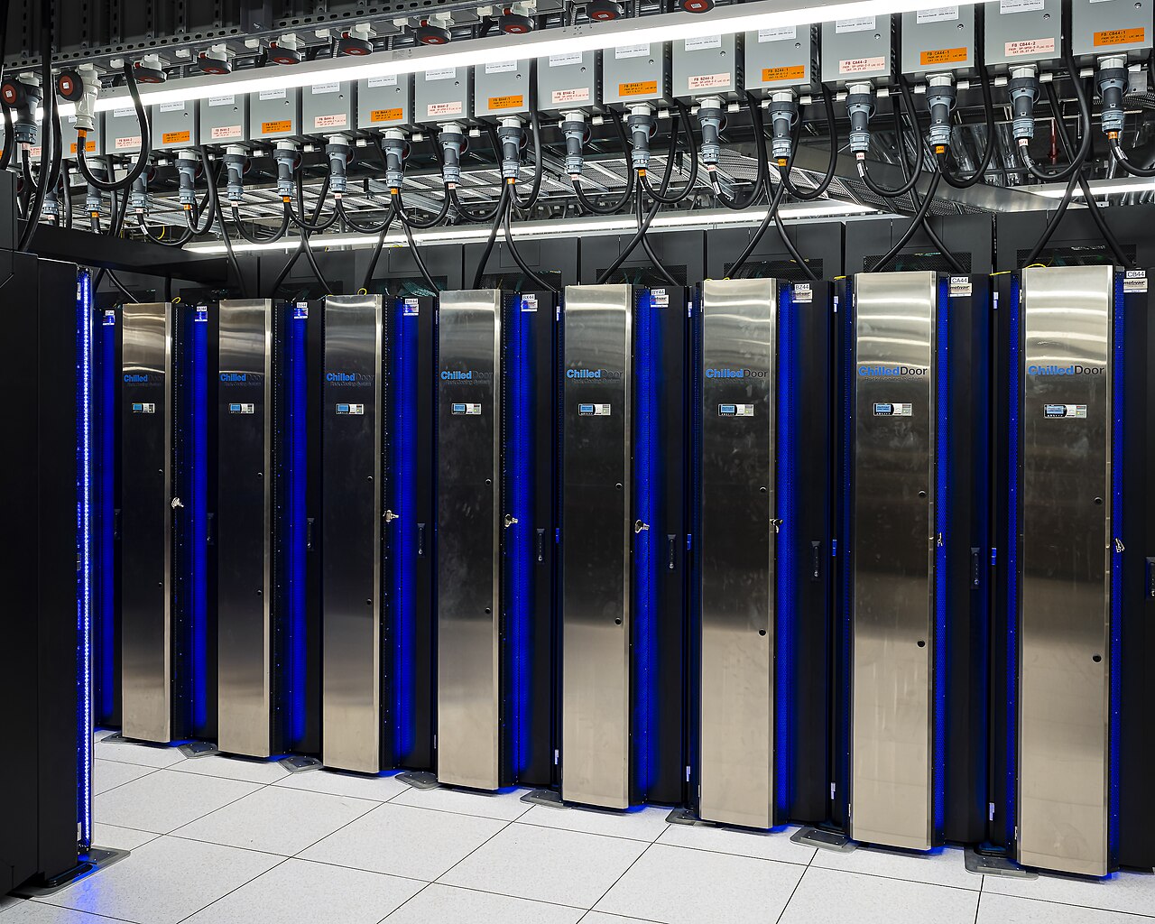 Fileira de gabinetes pretos e de aço inox com luzes azuis, do supercomputador Frontier, com cabos de energia presos ao teto.