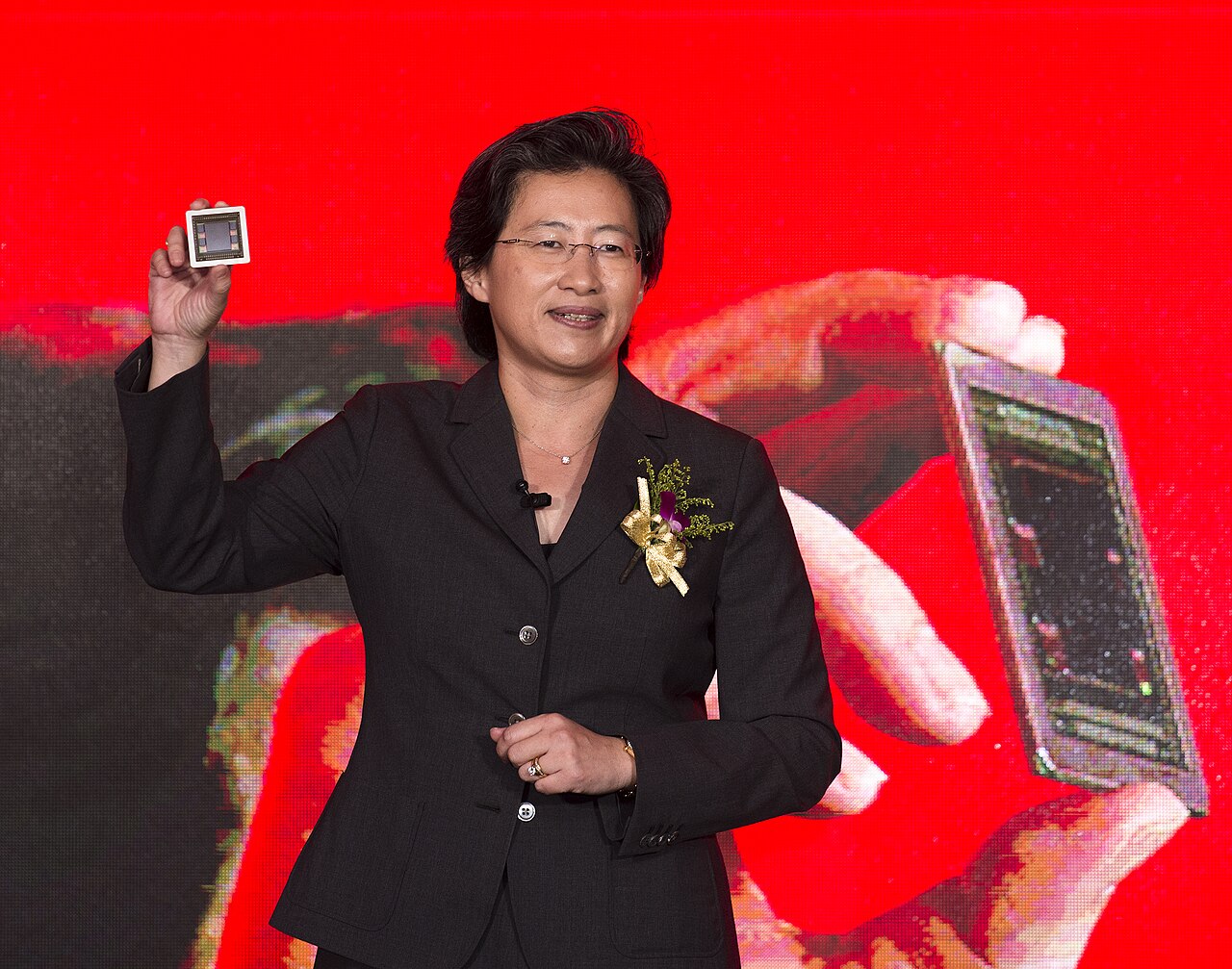 Lisa Su, de blazer preto com uma flor dourada na lapela, ergue um chip entre os dedos diante de um telão vermelho.