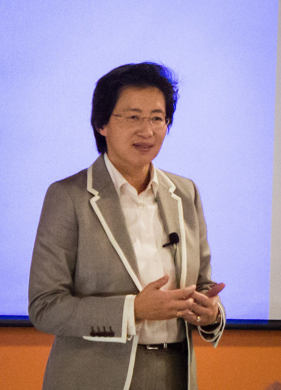 Lisa Su de blazer cinza com friso branco e camisa branca, falando de pé diante de uma tela de projeção azul clara.