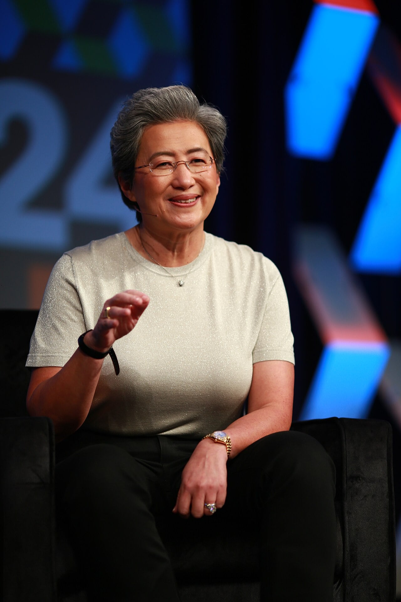 Lisa Su, de cabelo curto grisalho e óculos finos, sorri e gesticula sentada num palco, com telões coloridos ao fundo.