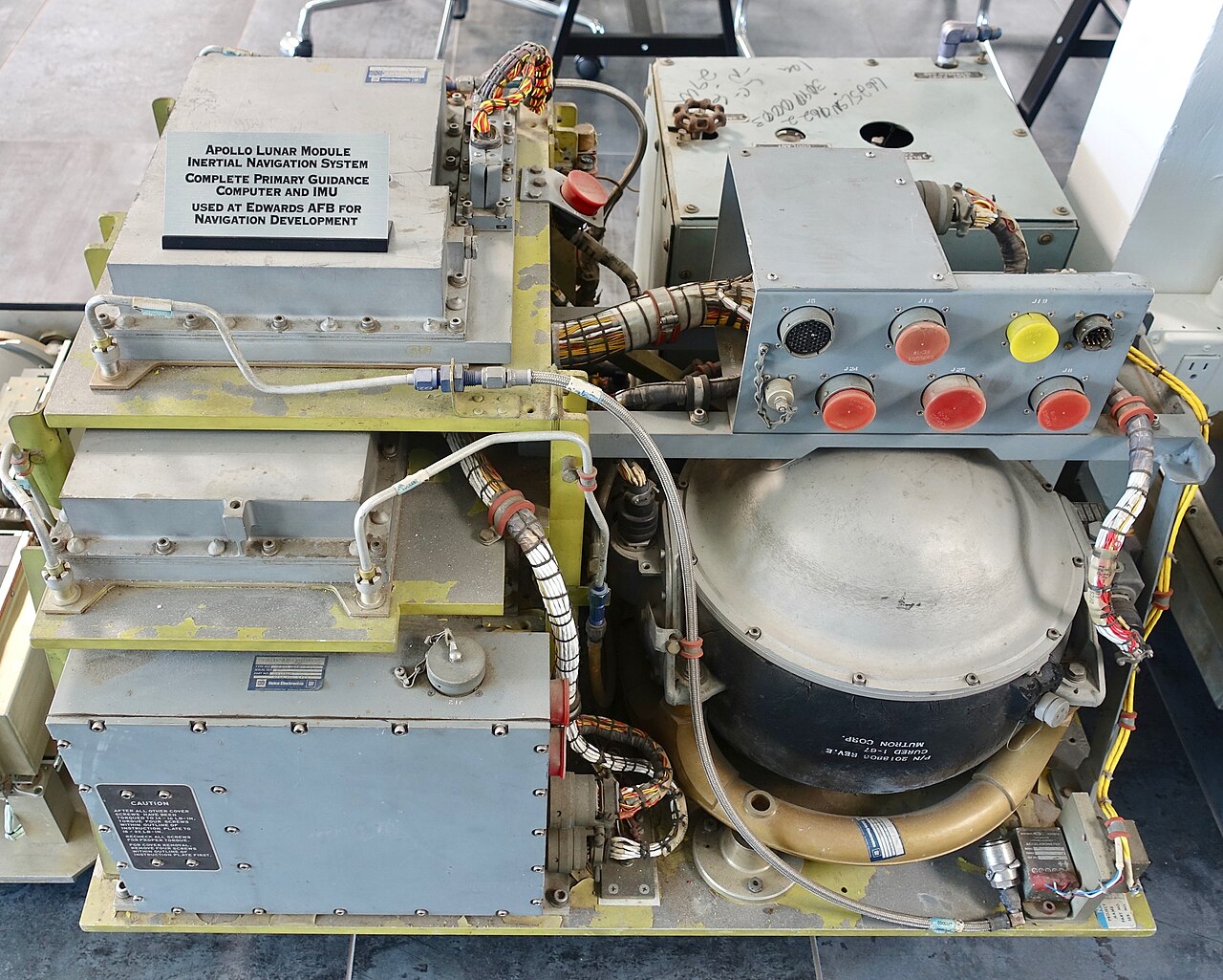 Conjunto de navegação inercial do módulo lunar visto de cima, com caixas metálicas, cabos e uma grande cúpula escura, sobre uma estrutura amarela e uma placa explicativa.