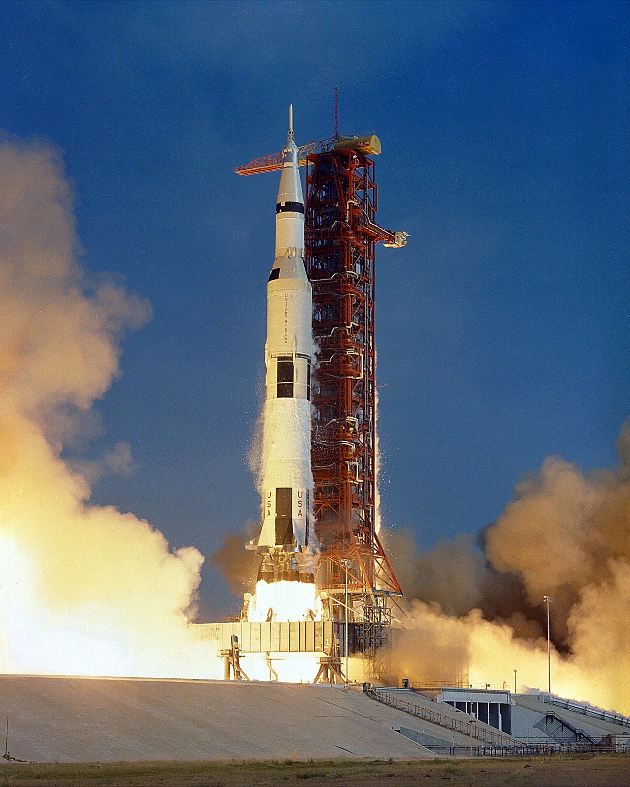 O foguete Saturn V da Apollo 11 sobe de sua plataforma em meio a fumaça e chamas, em tomada vertical.