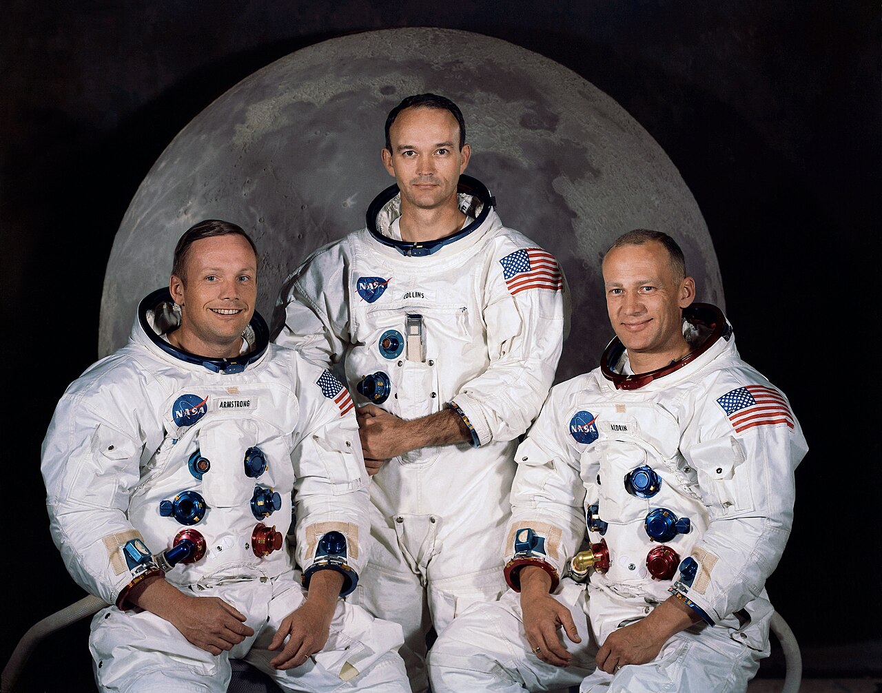 Retrato oficial dos três astronautas da Apollo 11, de traje espacial branco, sentados e de pé diante de uma imagem grande da Lua.
