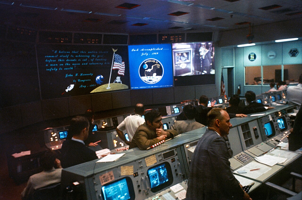 Sala de controle de missão em Houston, com fileiras de consoles e controladores sentados diante de telas que mostram o emblema da Apollo 11 e a frase Task Accomplished.