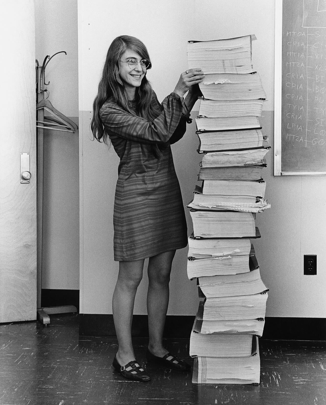 Fotografia em preto e branco de Margaret Hamilton, sorrindo, de pé ao lado de uma pilha de listagens de computador mais alta que ela.