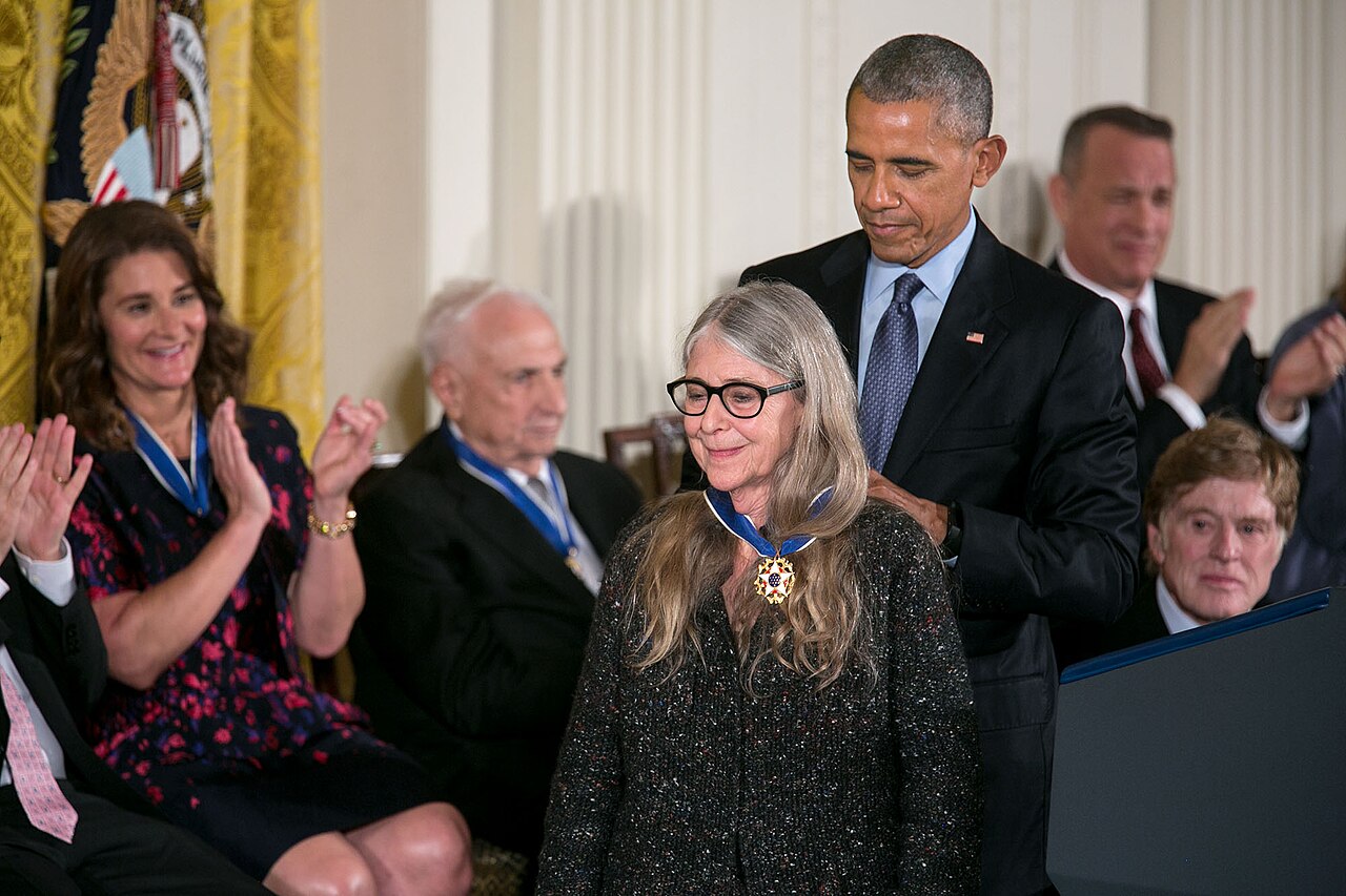 Barack Obama, de pé atrás de Margaret Hamilton, ajusta a fita azul da Medalha Presidencial da Liberdade no pescoço dela, enquanto convidados aplaudem ao fundo.