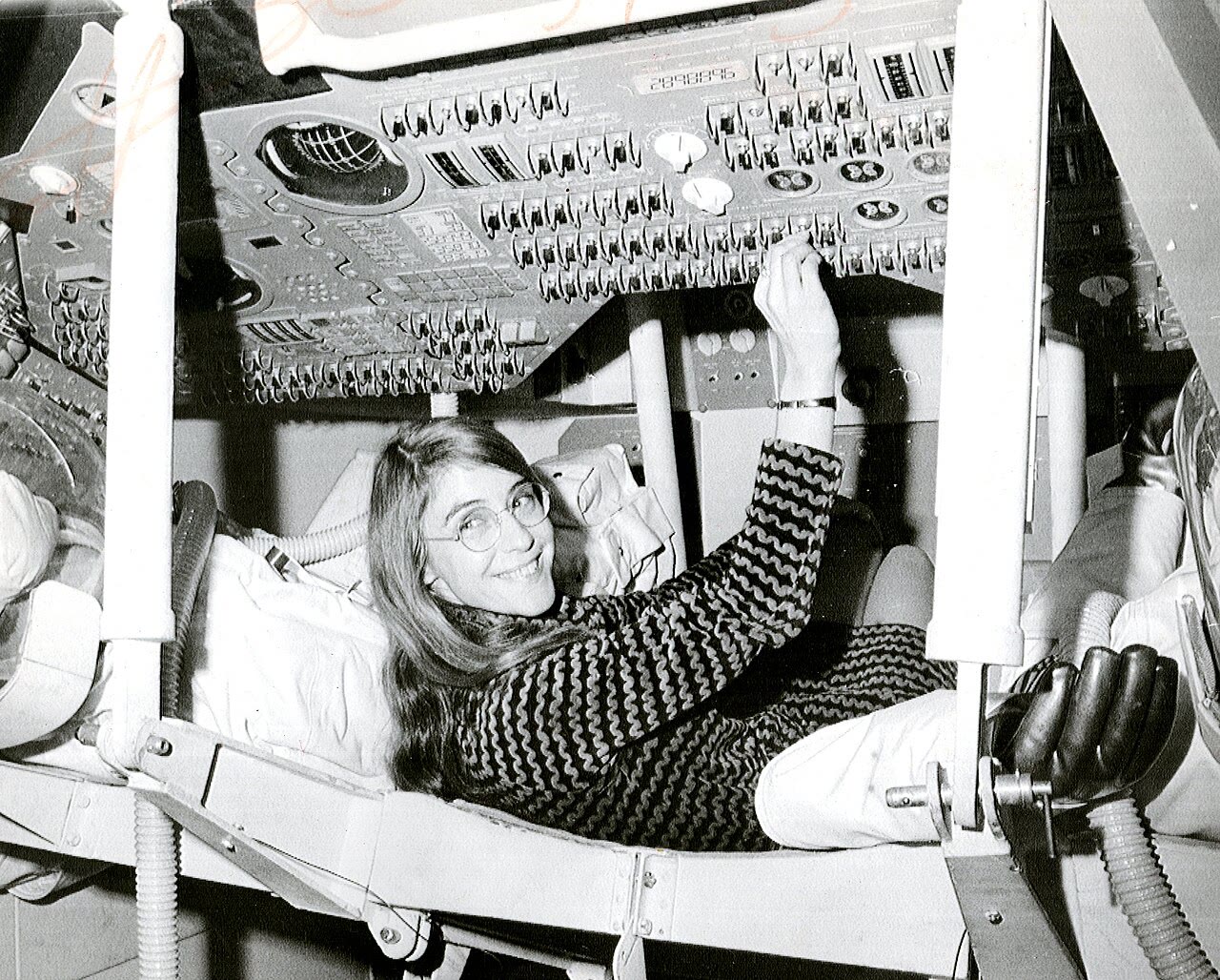 Margaret Hamilton, sorrindo, deitada num assento de cabine da Apollo e apoiando a mão num painel cheio de interruptores.