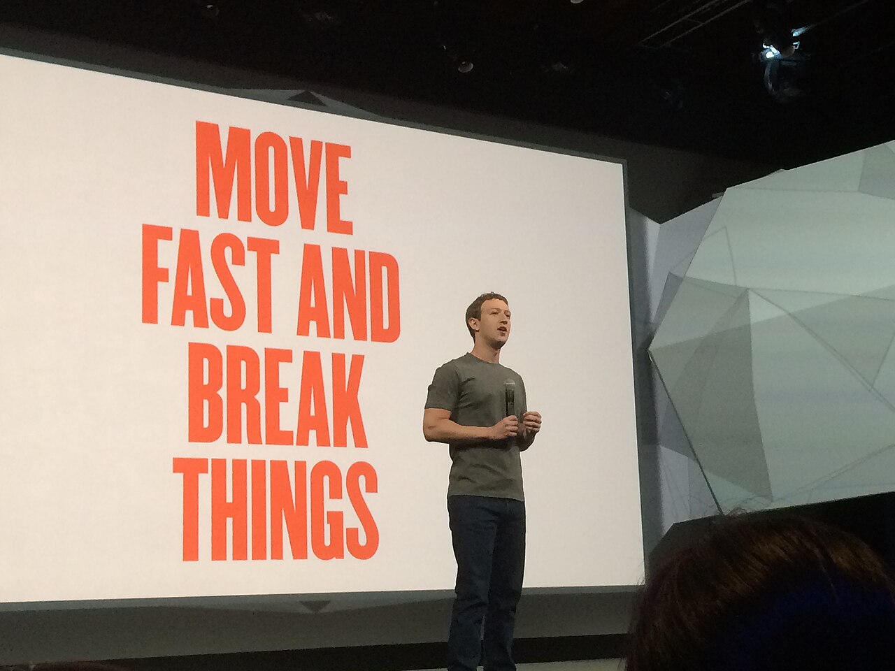 Zuckerberg, de camiseta cinza e microfone na mão, em pé em um palco, ao lado de um telão com o texto Move fast and break things.