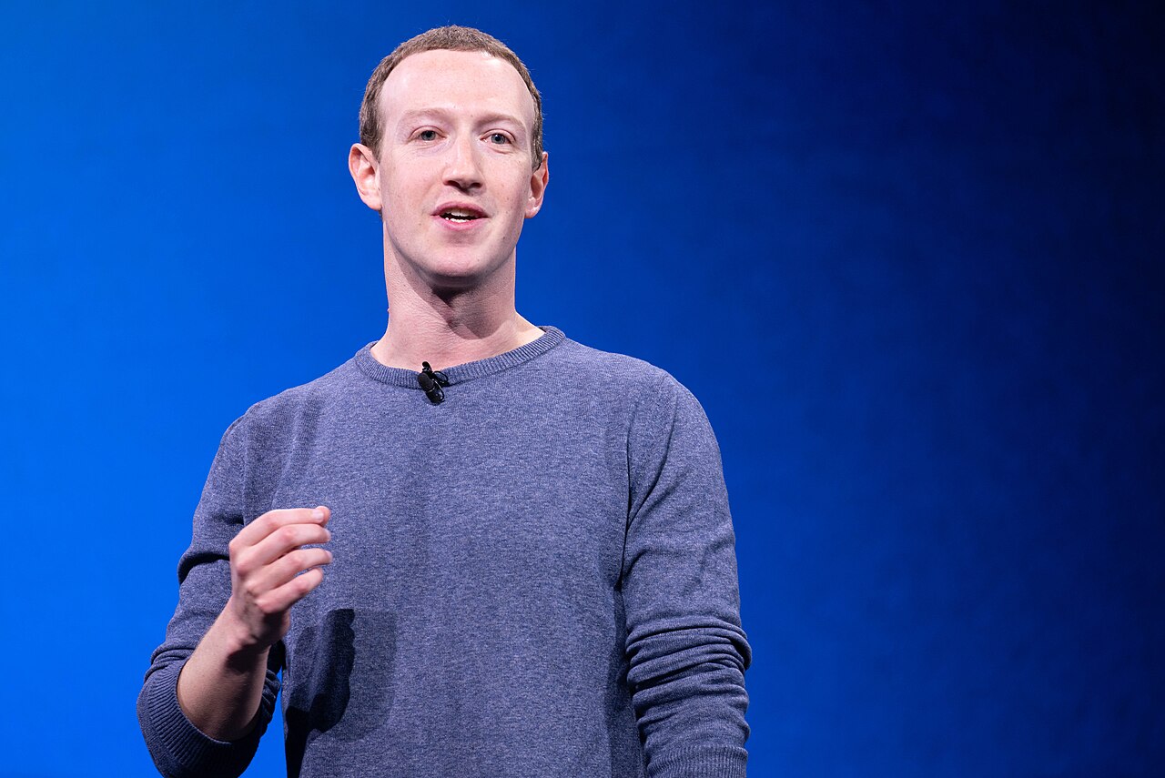 Mark Zuckerberg, de suéter azul-acinzentado e microfone de lapela, falando em um palco com fundo azul.