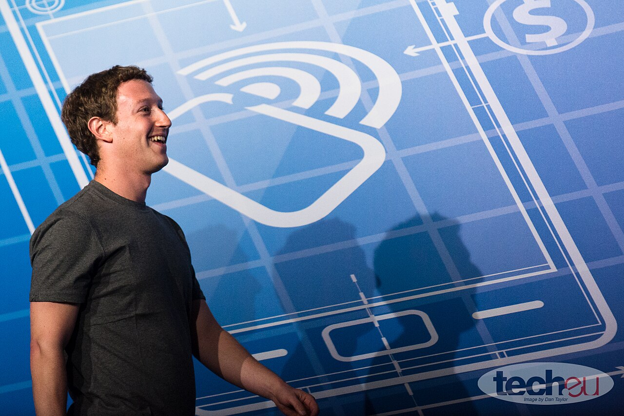 Mark Zuckerberg sorrindo, de camiseta cinza, diante de um painel azul com desenhos de um celular e um símbolo de sinal sem fio.