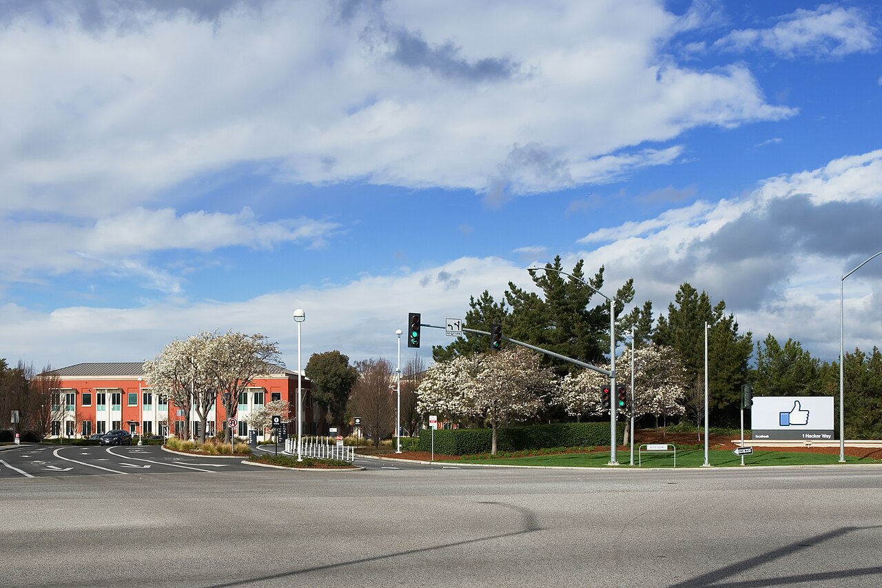 Entrada do complexo da sede do Facebook em Menlo Park, com a placa do polegar para cima e o endereço 1 Hacker Way, e um prédio vermelho ao fundo.