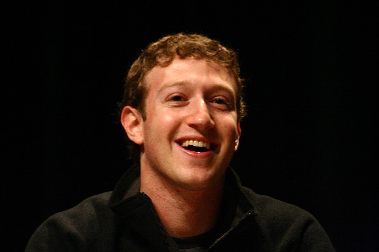 Mark Zuckerberg sorrindo, de cabelo cacheado e blusa escura de zíper, em foto de rosto sobre fundo preto.