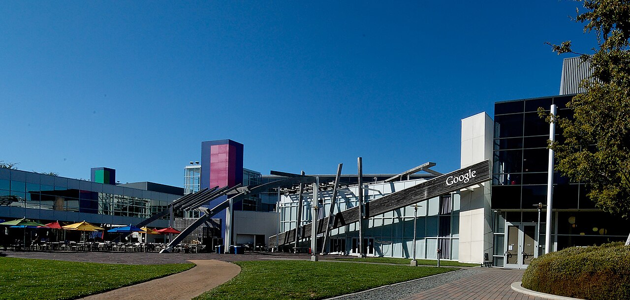 Pátio do campus do Google, com mesas e guarda-sóis coloridos, prédios de vidro e uma placa com a palavra Google em um telhado inclinado.
