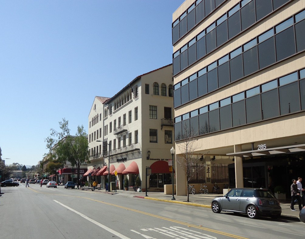 Rua do centro de Palo Alto com prédios de cores claras e toldos vermelhos de um lado e um edifício comercial moderno do outro, sob céu azul.