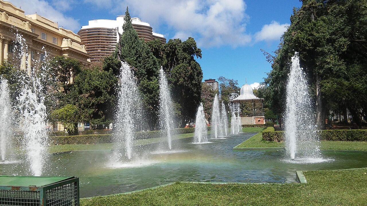 Chafarizes jorrando em um lago da Praça da Liberdade, em Belo Horizonte, com um pavilhão branco ao fundo e prédios entre árvores.