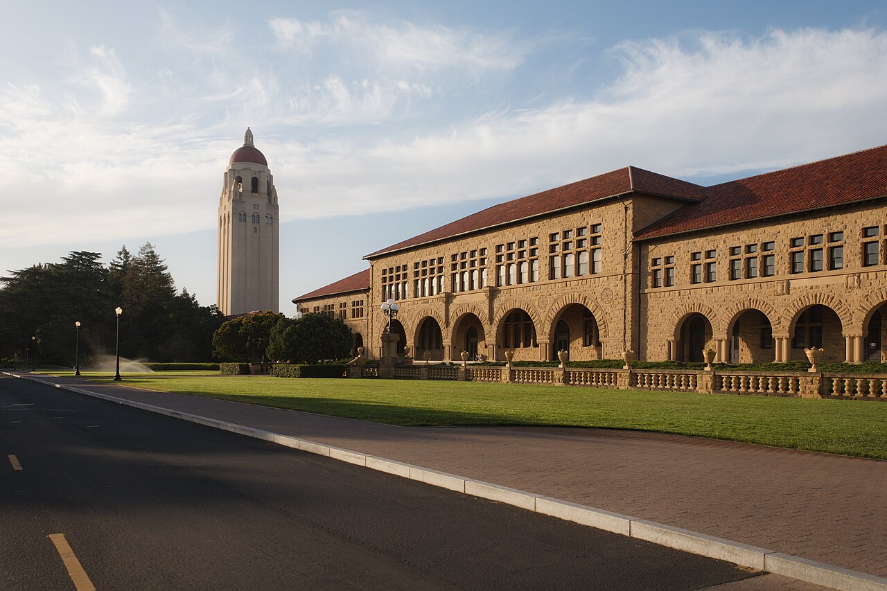 Torre Hoover, de cúpula vermelha, e um prédio de arenito com arcadas e telhado vermelho no campus de Stanford, sob céu azul com nuvens.