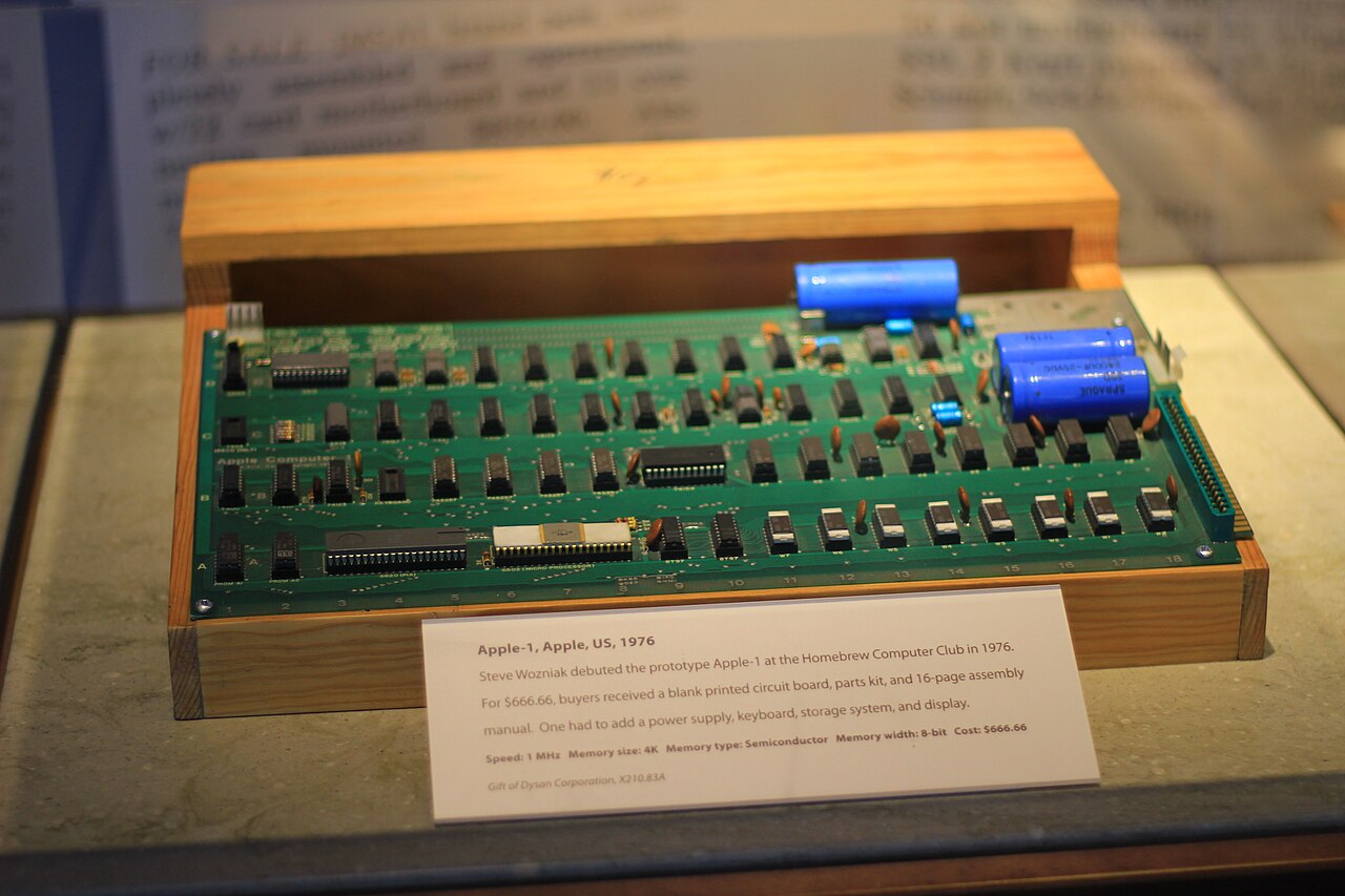 Placa de circuito verde do Apple-1, com fileiras de chips e capacitores azuis, sobre uma base de madeira, com a etiqueta do museu à frente.