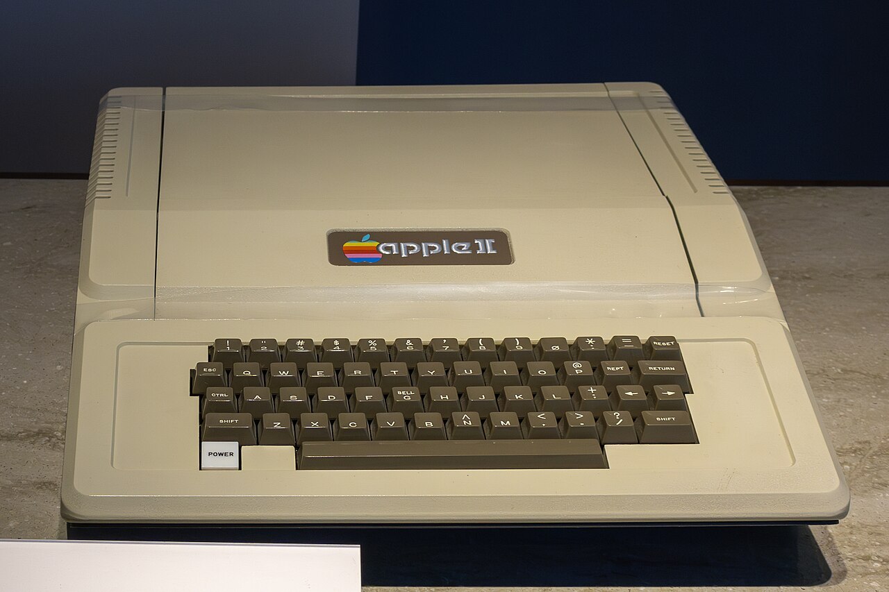 Apple II bege de 1977, com o logotipo colorido da maçã e o teclado integrado na frente, exposto sobre uma base cinza.