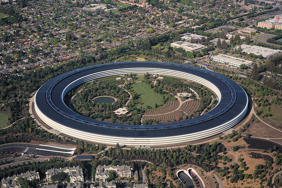 Vista aérea de um edifício circular enorme, com telhado coberto de painéis solares e áreas verdes, cercado por estacionamentos e ruas.