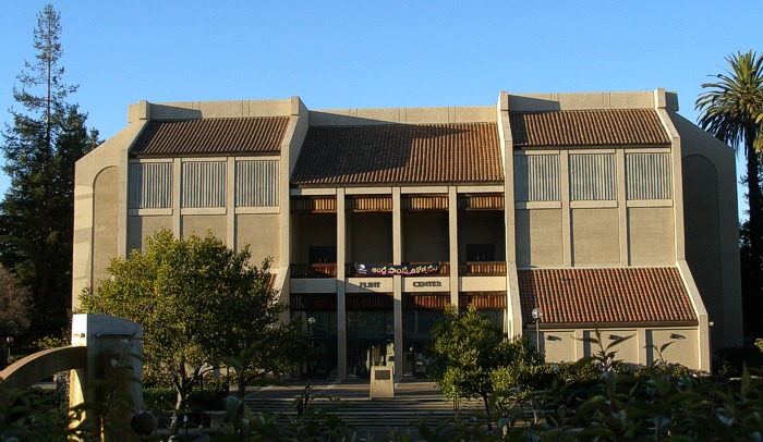 Fachada de concreto bege do Flint Center, com telhados de telhas vermelhas e o nome do auditório sobre a entrada, entre árvores.