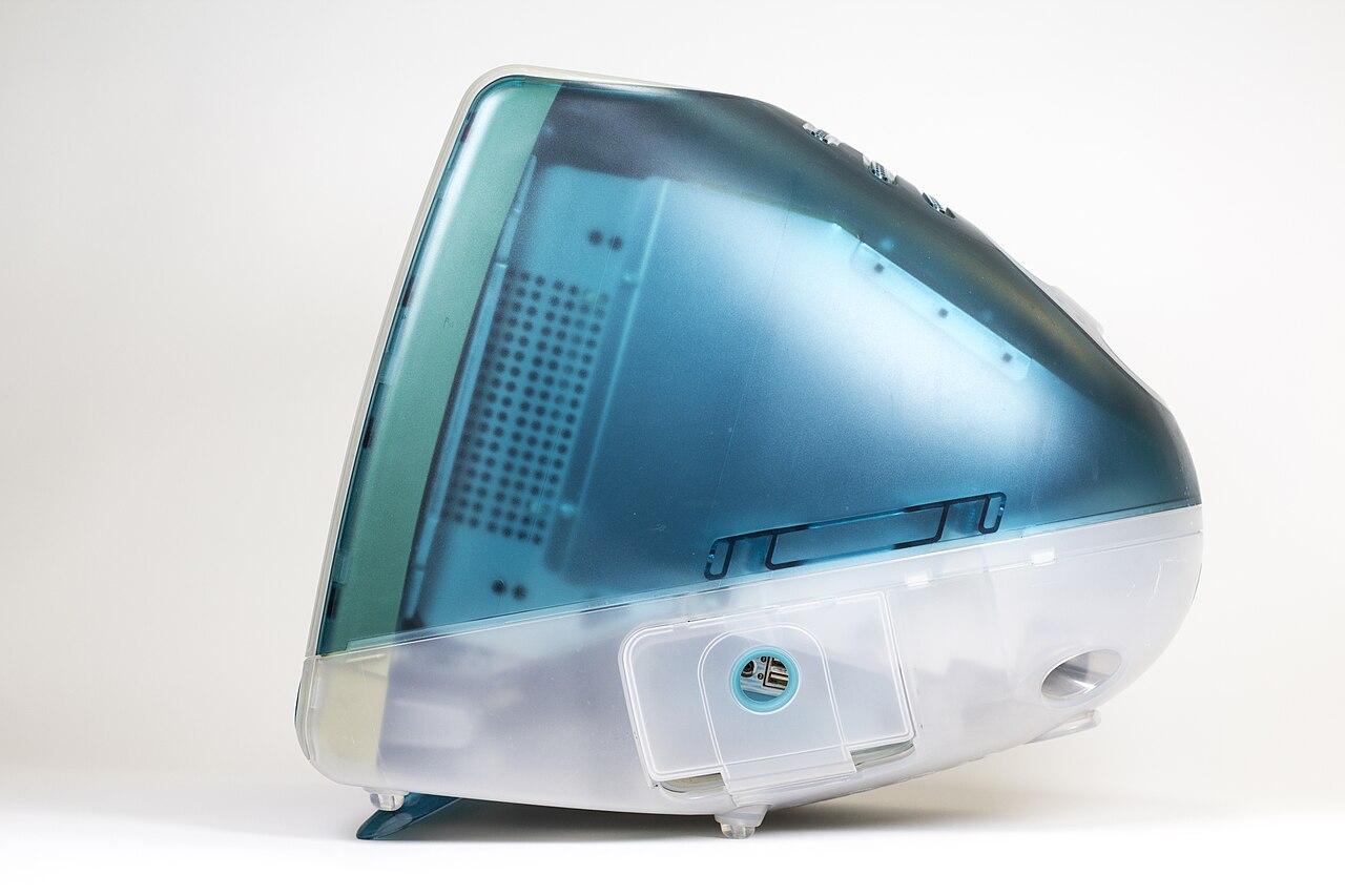 Vista lateral de um iMac G3 original azul-esverdeado e translúcido, com monitor de tubo e uma alça na parte de cima.