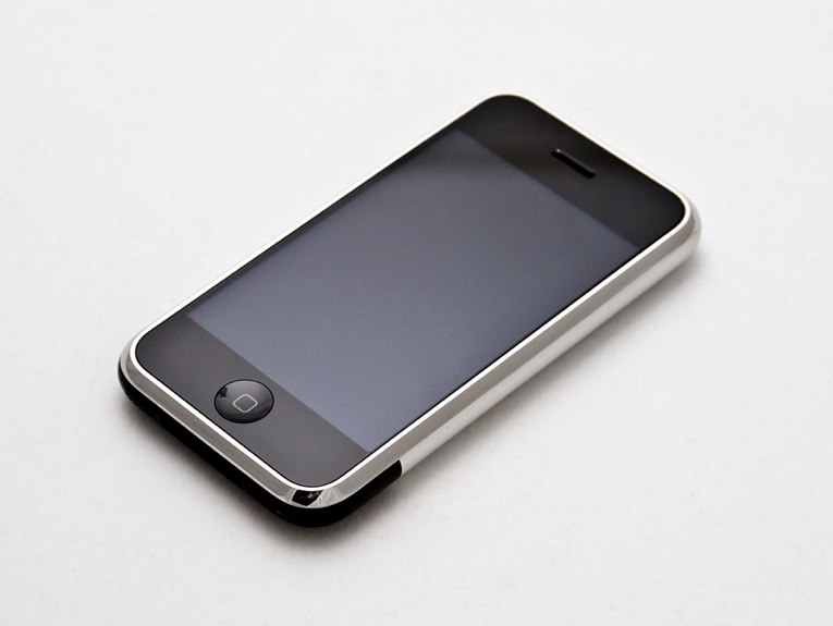 iPhone de primeira geração, com moldura prateada e botão redondo na parte de baixo, visto de cima em ângulo sobre fundo branco.
