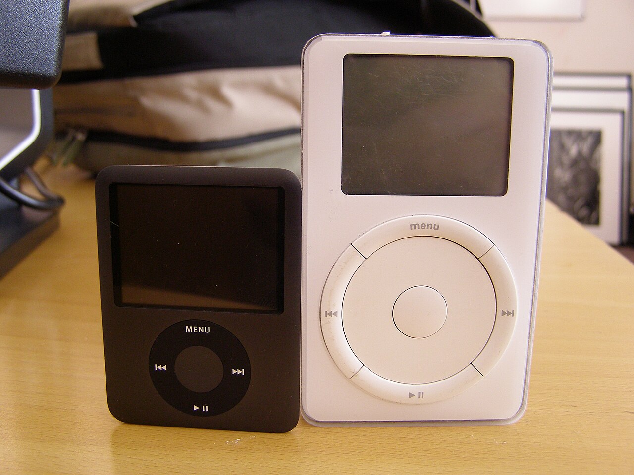 Dois tocadores lado a lado sobre uma mesa: um iPod nano preto, menor, à esquerda, e o iPod original branco de 5 GB, maior, com roda de controle, à direita.